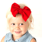 A toddler girl wearing an oversized red bow headband.  She is sitting in front of a white backdrop looking at the camera.