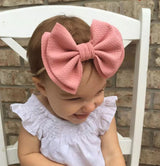 A baby girl sitting on a white wooden chair in front of a brown brick wall and wearing an oversized pink cotton bow around her head.  She is smiling and looking down.