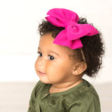 A baby girl wearing an oversized hot pink cotton bow around her head and sitting in front of a white backdrop.  She is smiling and looking to the left.