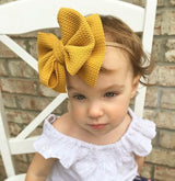 A baby girl sitting on a white wooden chair in front of a brown brick wall and wearing an oversized mustard coloured cotton bow around her head.  The bow has a chevron texture with a tan elastic.